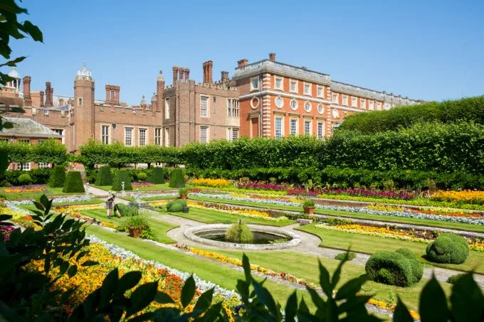 Manecured lawns of Hampton Court Palace with hues of green, yellow and red sit below Hampton Court Palace in the background and under a blue sky. Foliage frames the image.