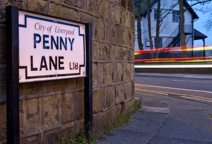 Street sign in white and black resting on a footpath with the words City of Liverpool - Penny Lane, with brick wall behind.