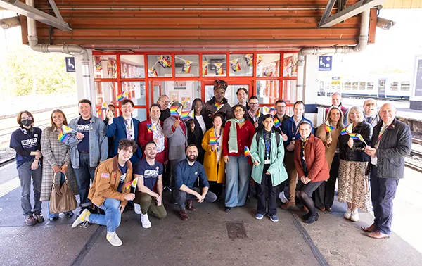 A large group of people, of different races, genders and ages, some holding rainbow flags, all facing the camera and smiling while on a station platform. 