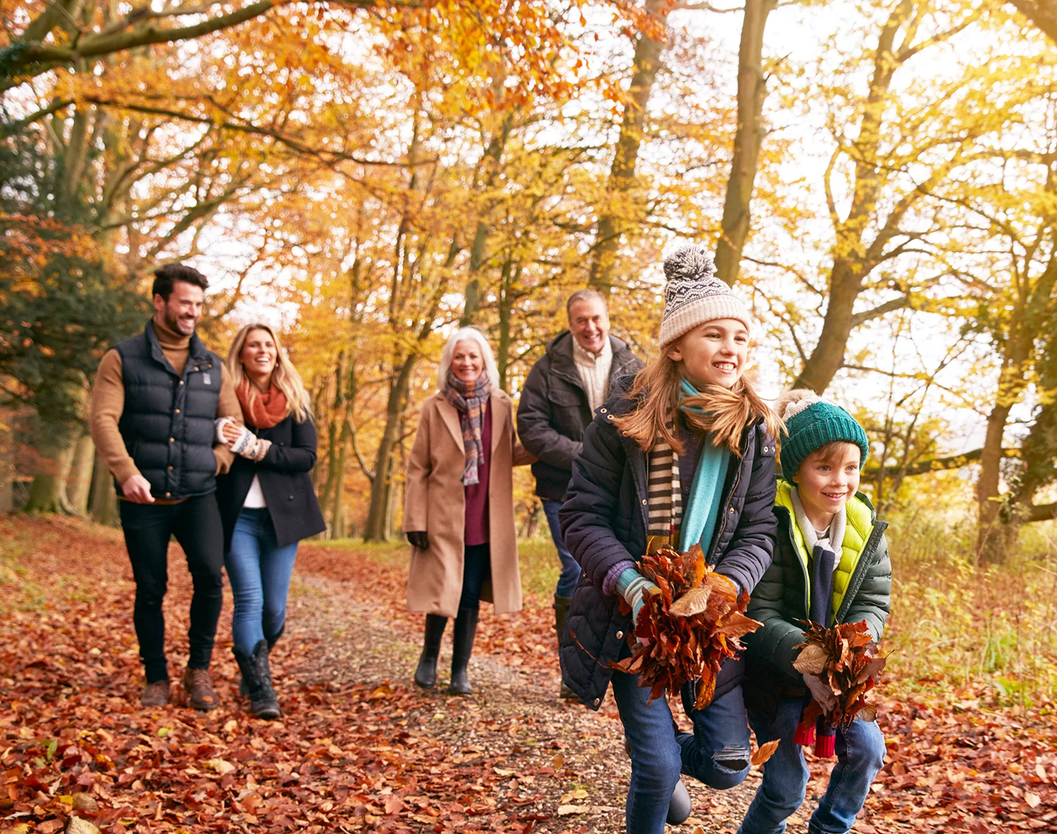 A multigenerational white family dressed in warm clothing, walking among fallen leaves in a wood on an autumn day. 