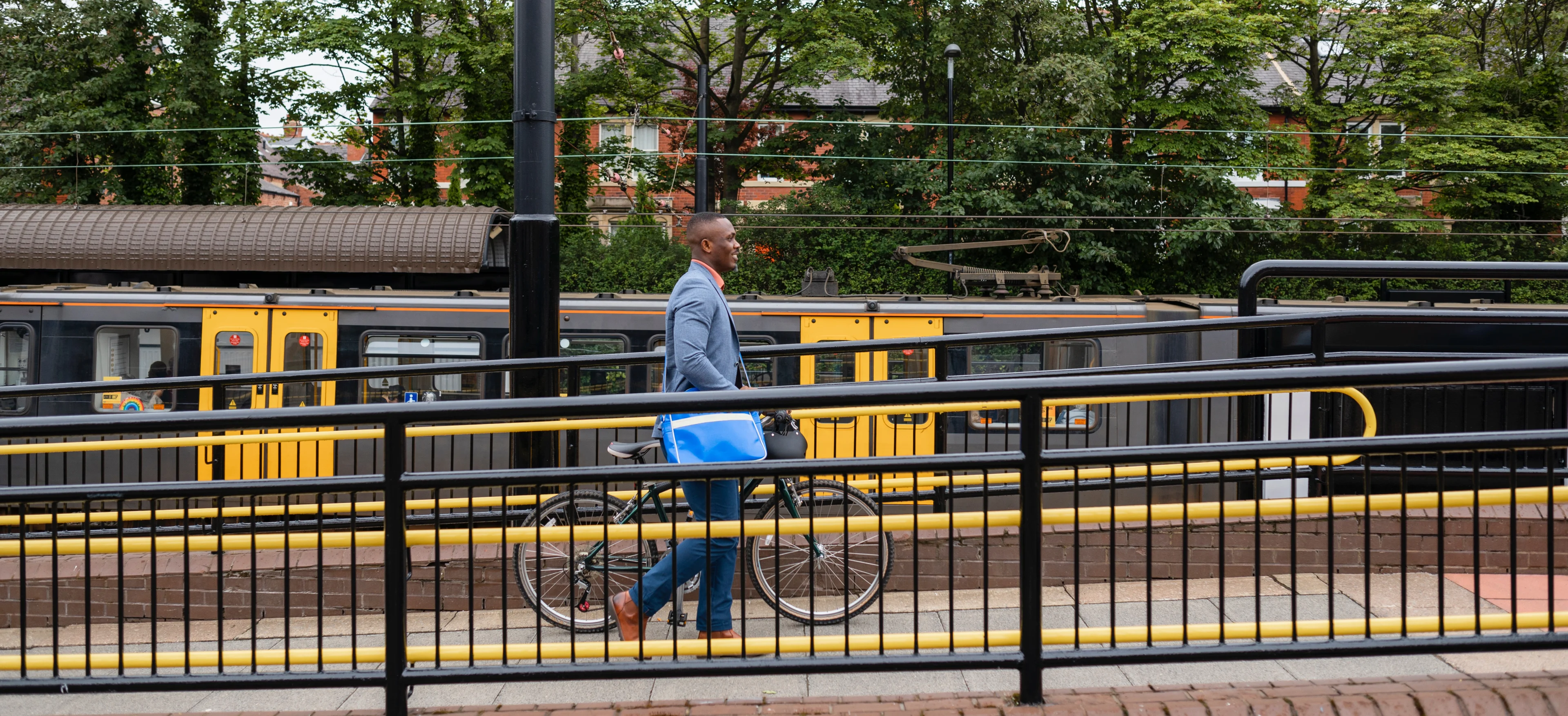 A man pushes a bicycle up a ramp at a train station, with a train at the platform in the background