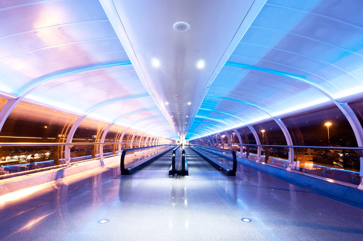 Moving walkway at Manchester Airport illuminated by blue lighting in a curved glass tunnel at night.