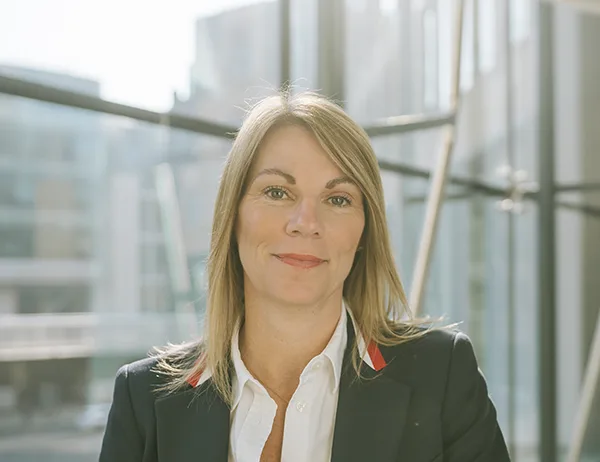 A white woman with long straight blonde hair looking at the camera, she is wearing a white shirt and black jacket. 