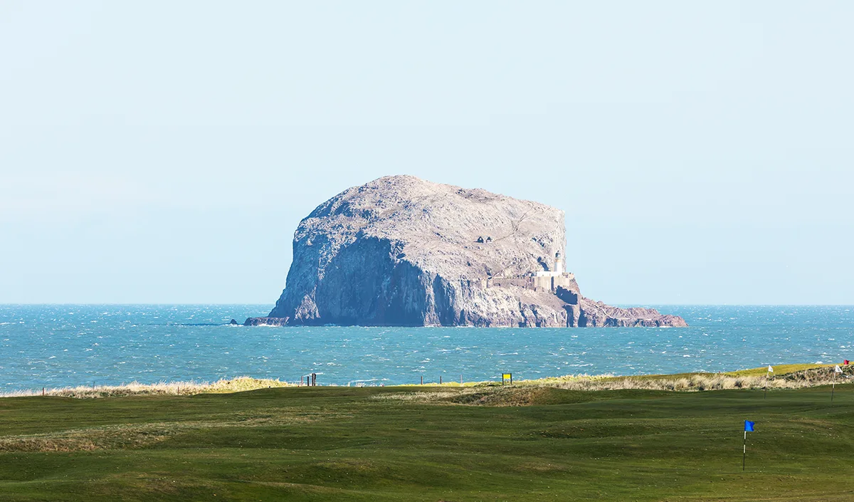 A rocky outcrop in the sea, near to the shore. A golf course runs up to the water. 