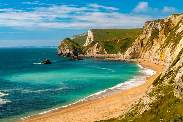 A sandy beach with clear turquoise sea and chalky cliffs above, on a sunny day.