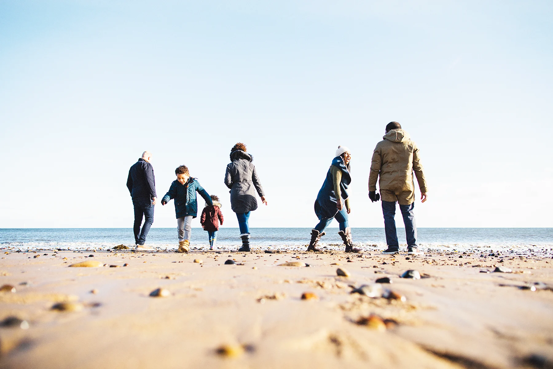 A multigenerational mixed-race family standing at the edge of the sea on a beach on a cold but sunny day.