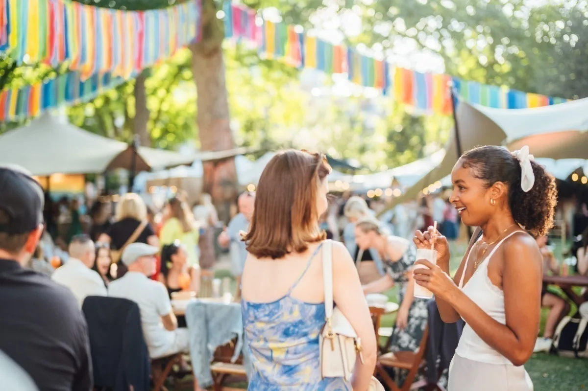A young white woman and a young Black woman chatting and enjoying a drink at a busy outdoor event on a sunny day, with bunting and marquees in the background.