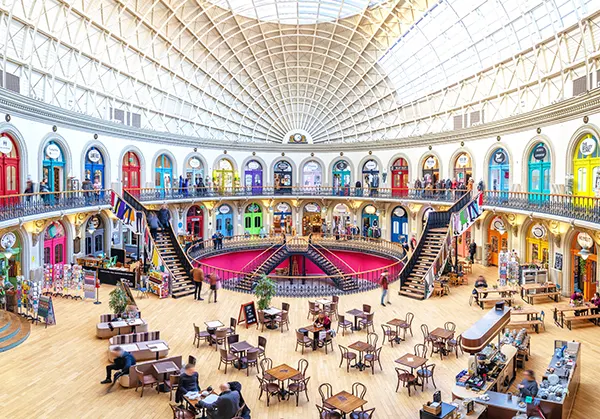The interior of a circular shopping centre building with cafe tables on a mezzanine floor and a variety of brightly coloured doors on 2 levels. 