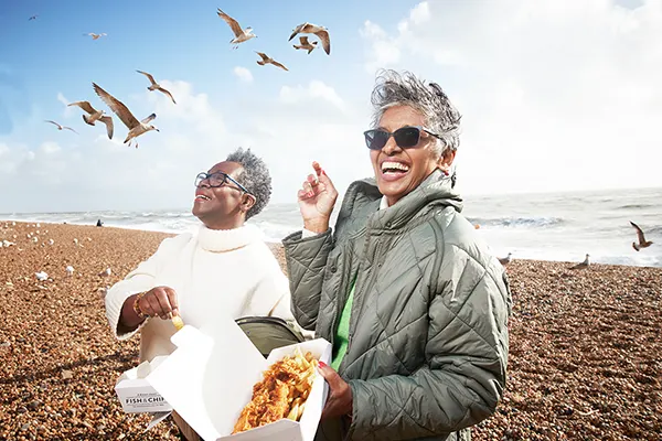 Two Black women laughing and eating fish and chips on a pebble beach while seagulls swoop around them. 