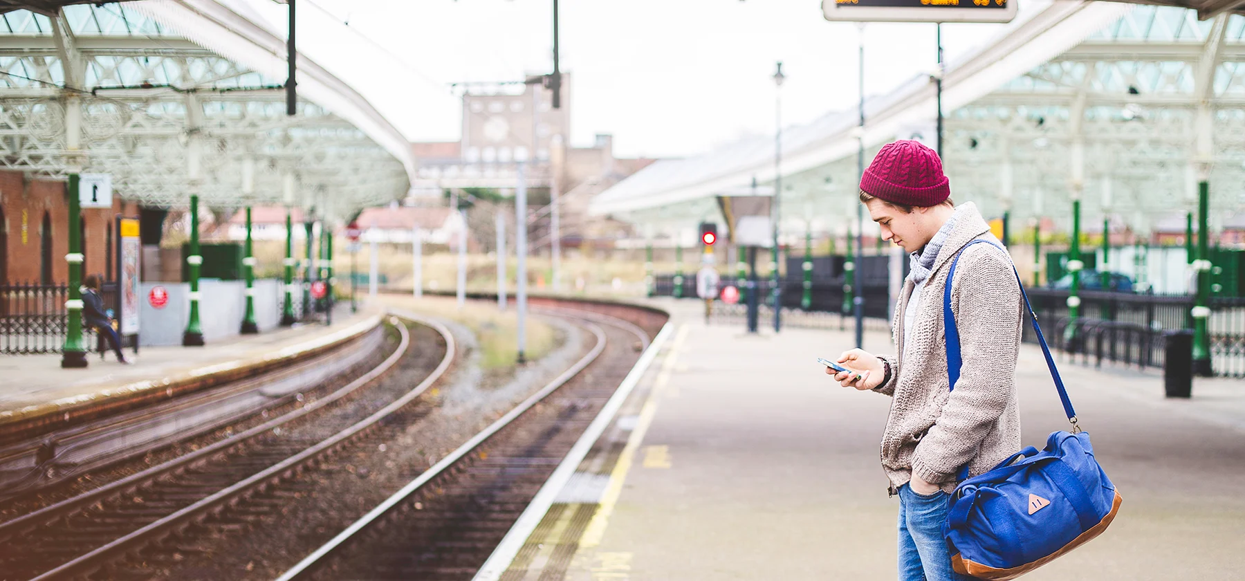 A young man wearing a red wooly hat stands looking at his phone on an empty station platform during the day.