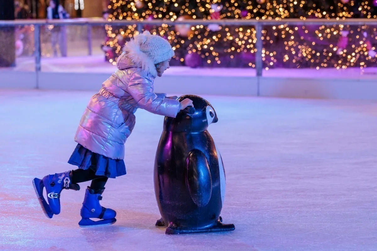A young white girl in a wooly hat and winter coat, wearing blue ice skates, pushes a penguin-shaped skating helper on an ice rick