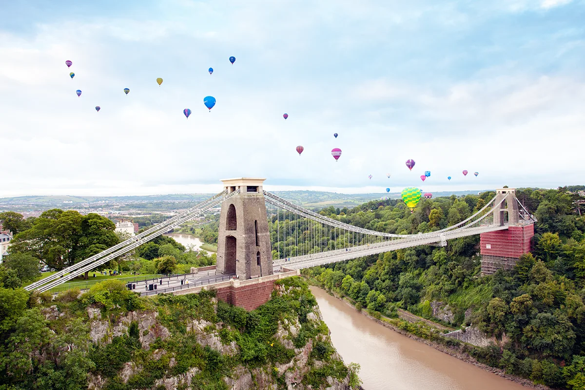 An aerial shot of a suspension bridge over a river, with hills and trees on either side, and a number of hot air balloons floating above in a blue sky.