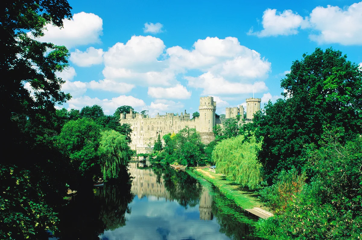 An old English light stone castle with towers and battlements, set in woodlands next to a river.