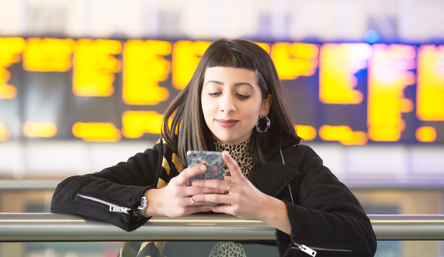 A woman with olive skin and long dark hair with a blunt fringe looks at her mobile phone screen while standing in front of a large train station departure board.