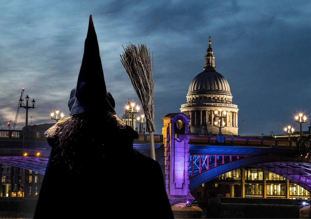 Back view of a woman in a witches hat and cape, holding a broomstick, and looking across the Thames towards St Pauls Cathedral at twilight. 