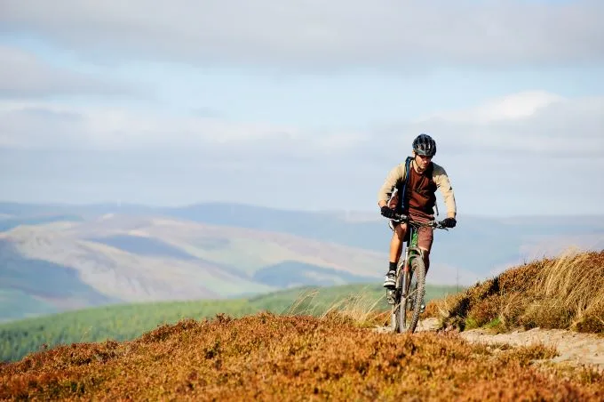 Man cycles up a hilly pathway with brambles lining either side. Mountains sit hazily in the background under a cloudy and blue sky.
