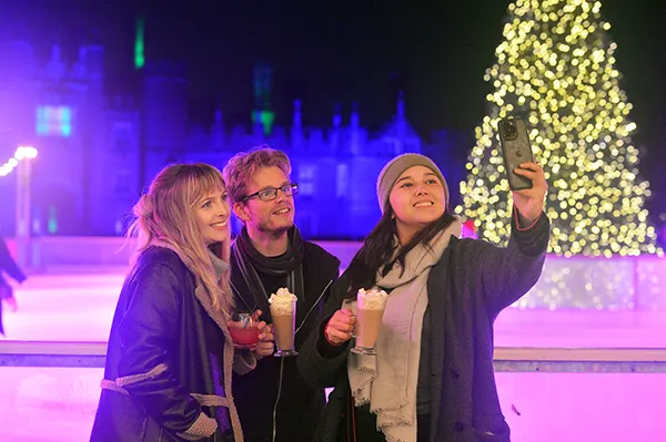 2 white women and a white man standing on at outdoor ice rink and taking a selfie in front of a palace with a large Christmas tree covered in lights.