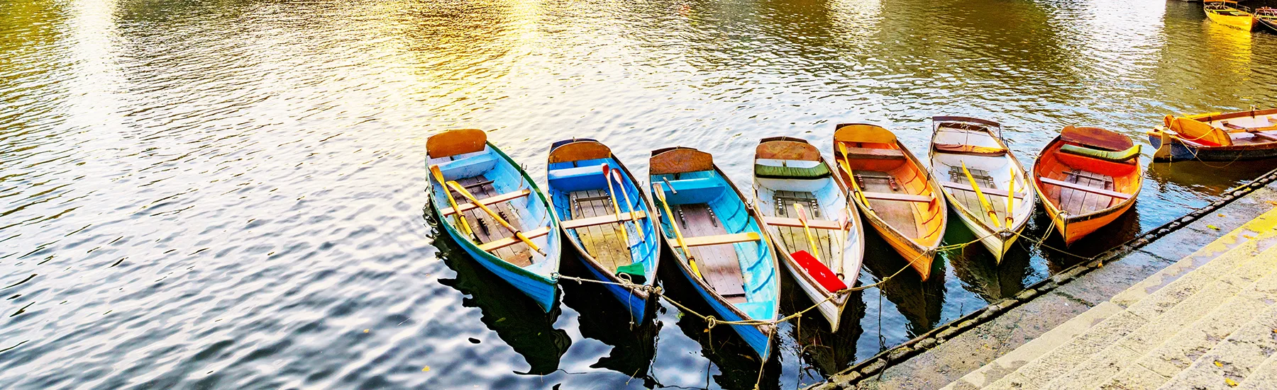 Colourful rowing boats moored on a river with a low stone bridge in the background. 