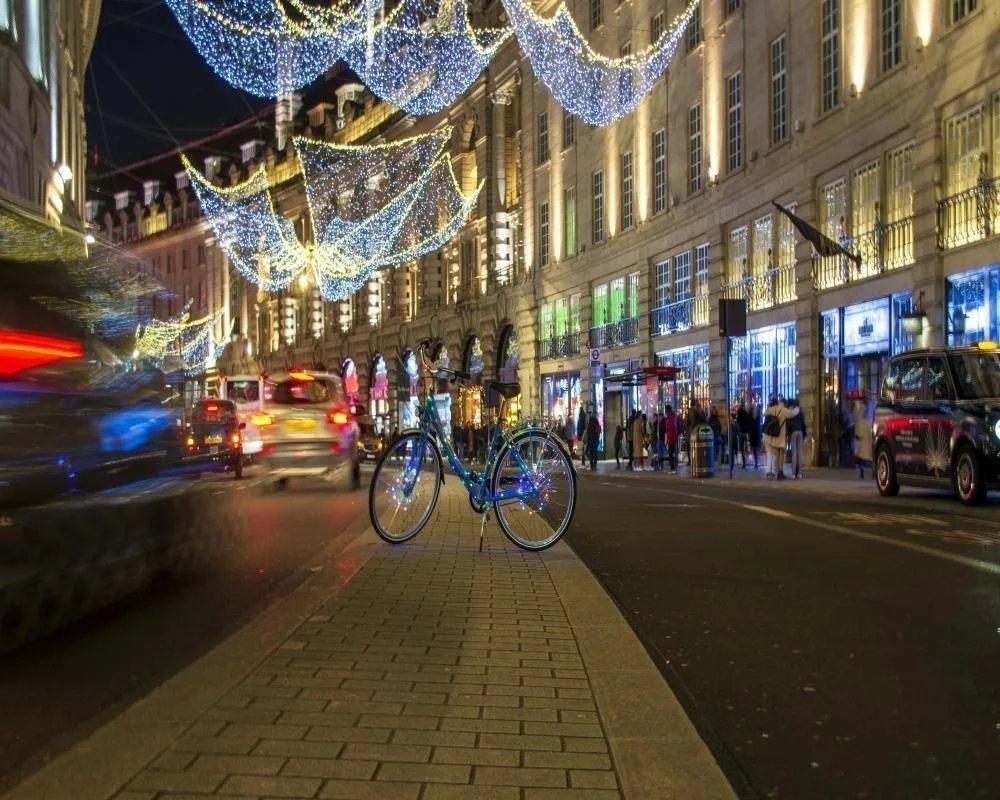 Illuminated bicycle on a London street at night with festive blue and gold Christmas lights hanging above shops.