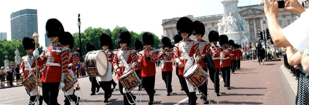 Royal Guards marching in ceremonial red uniforms with bearskin hats outside Buckingham Palace during the Changing of the Guard.