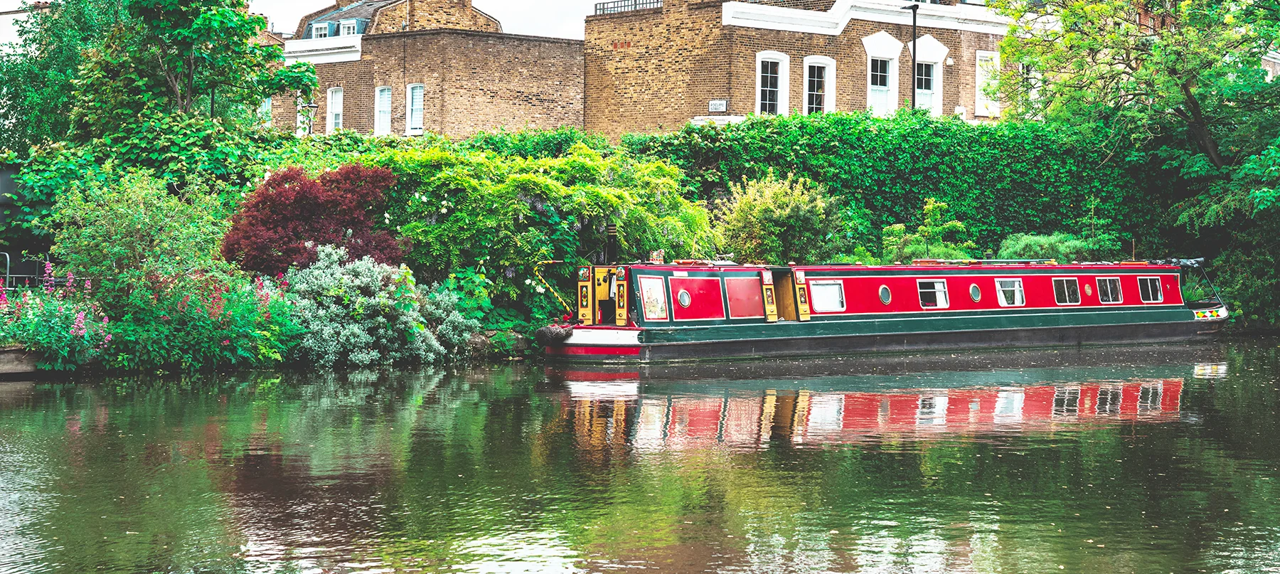 A red barge moored on a canal surrounded by greenery with brick houses in the background. 