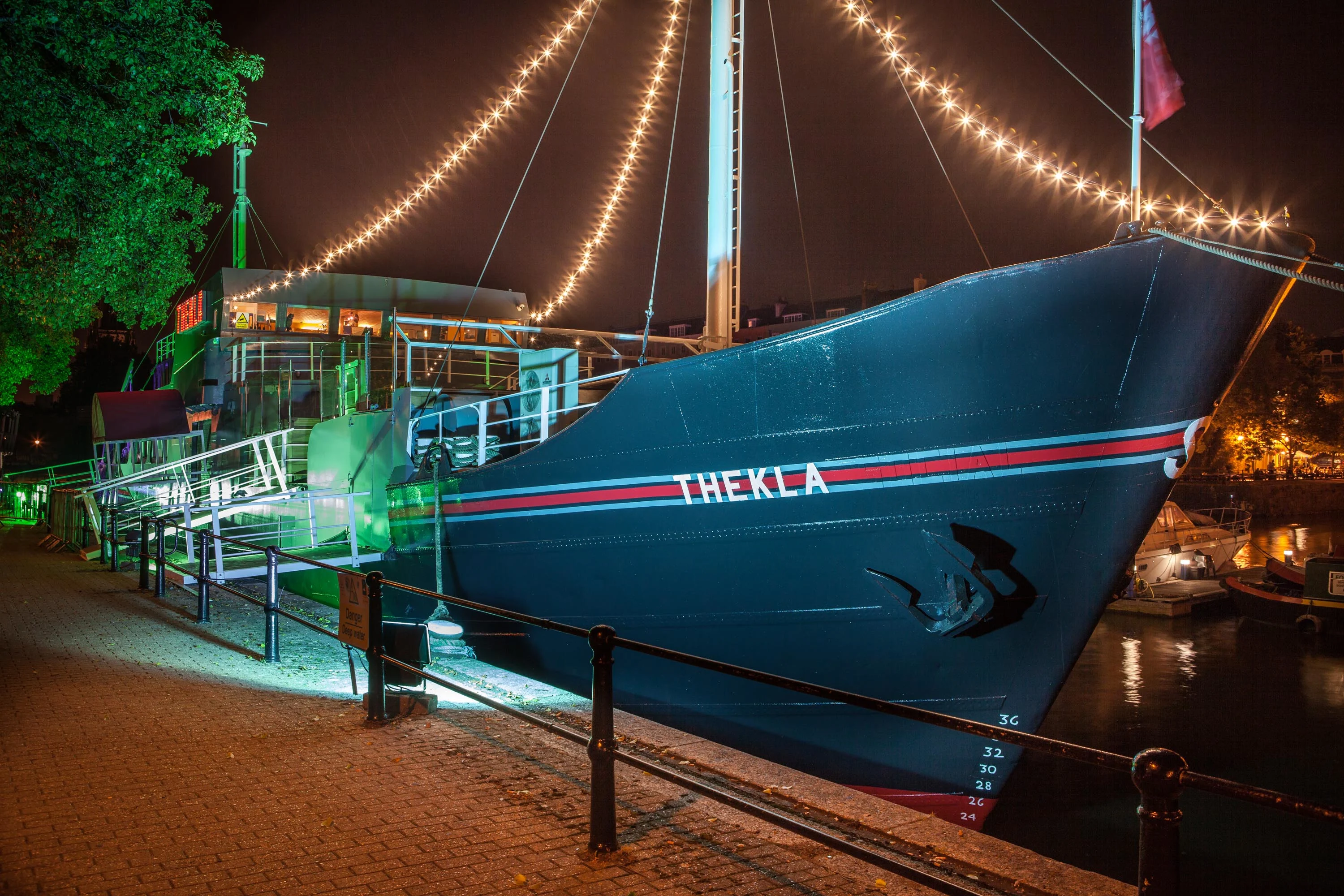 A docked boat with the name THEKLA painted on the side and strings of lights hanging from the mast. 