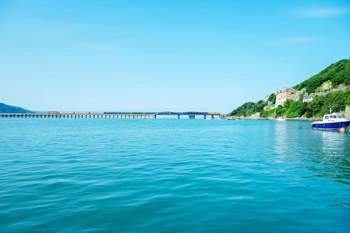 A train crossing a bridge over a bright blue sea, with a boat and tree-lined cliffs to the right. 