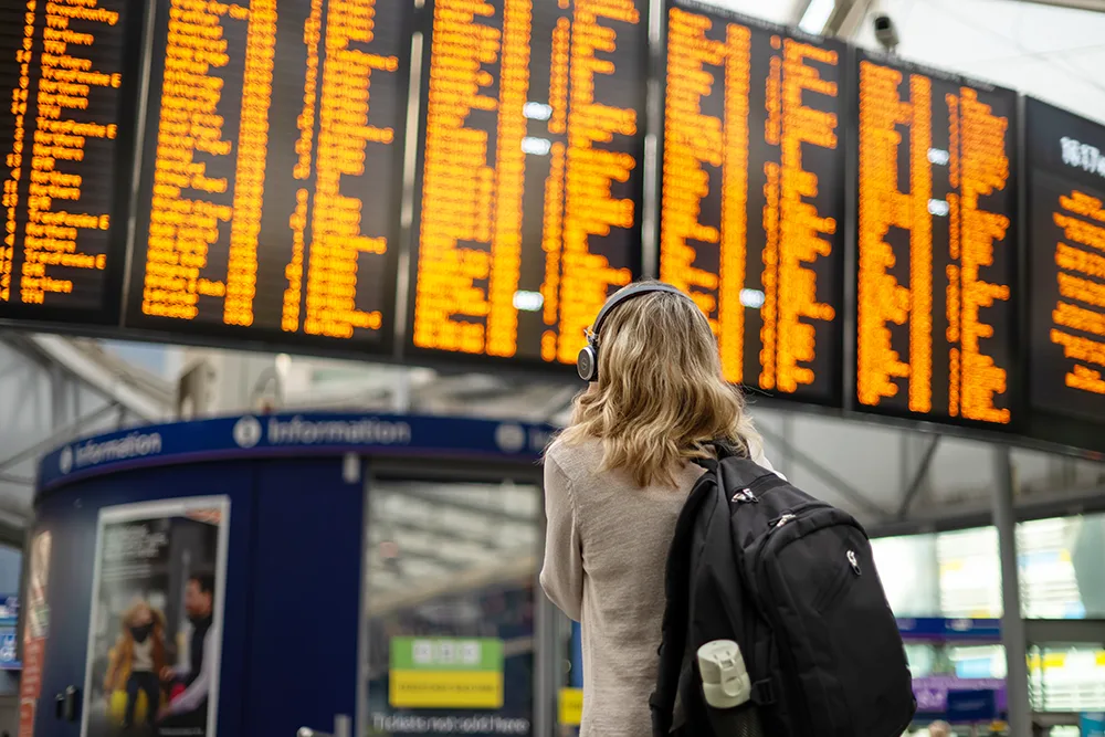 Back view of a woman with long blonde hair carrying a black rucksack, standing at a station and looking up at the large electronic departure board.
