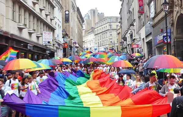 A large crowd of people at an LGBTQIA+ Pride parade, they are holding a huge rainbow flag horizontally between them, and many people are carrying rainbow umbrellas.