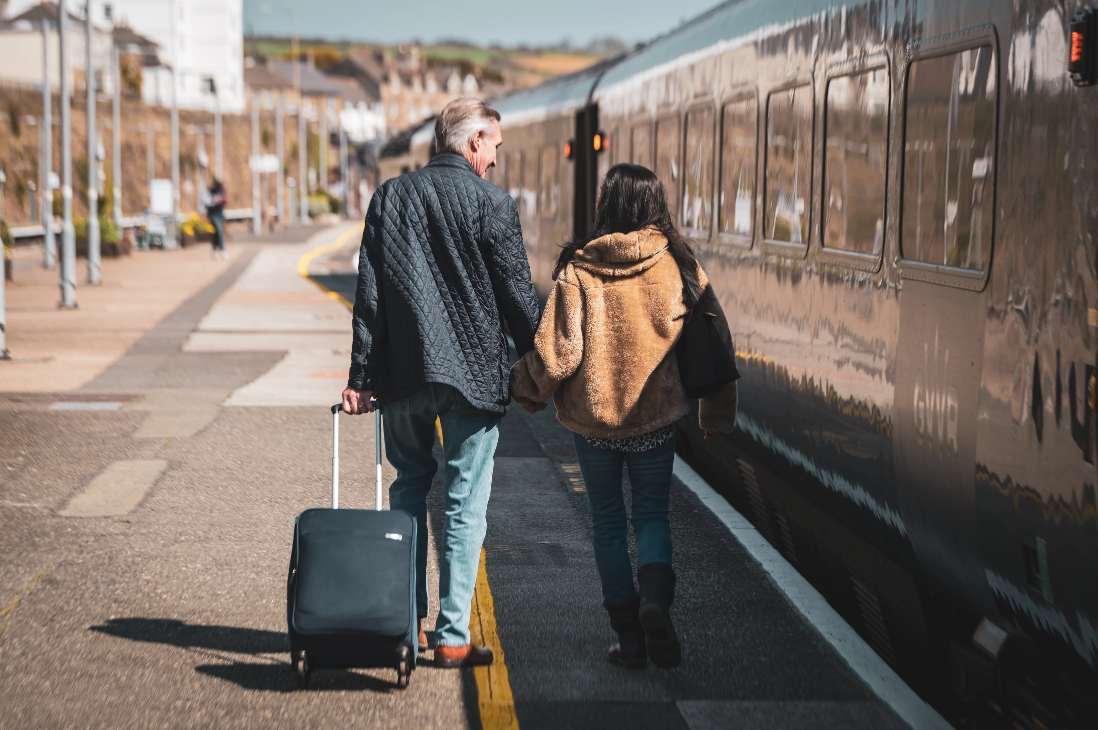 A man and a woman seen from behind holding hands and walking down a railway station platform in the daytime