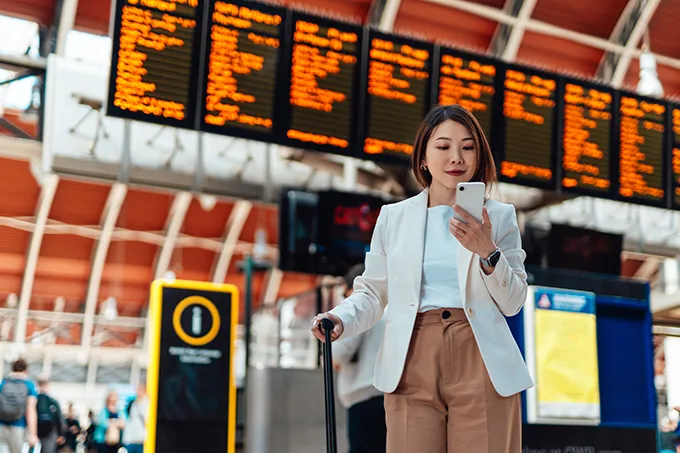 An East Asian woman standing in front of a large departure board at a rail station, looking at her phone and holding a wheeled suitcase.