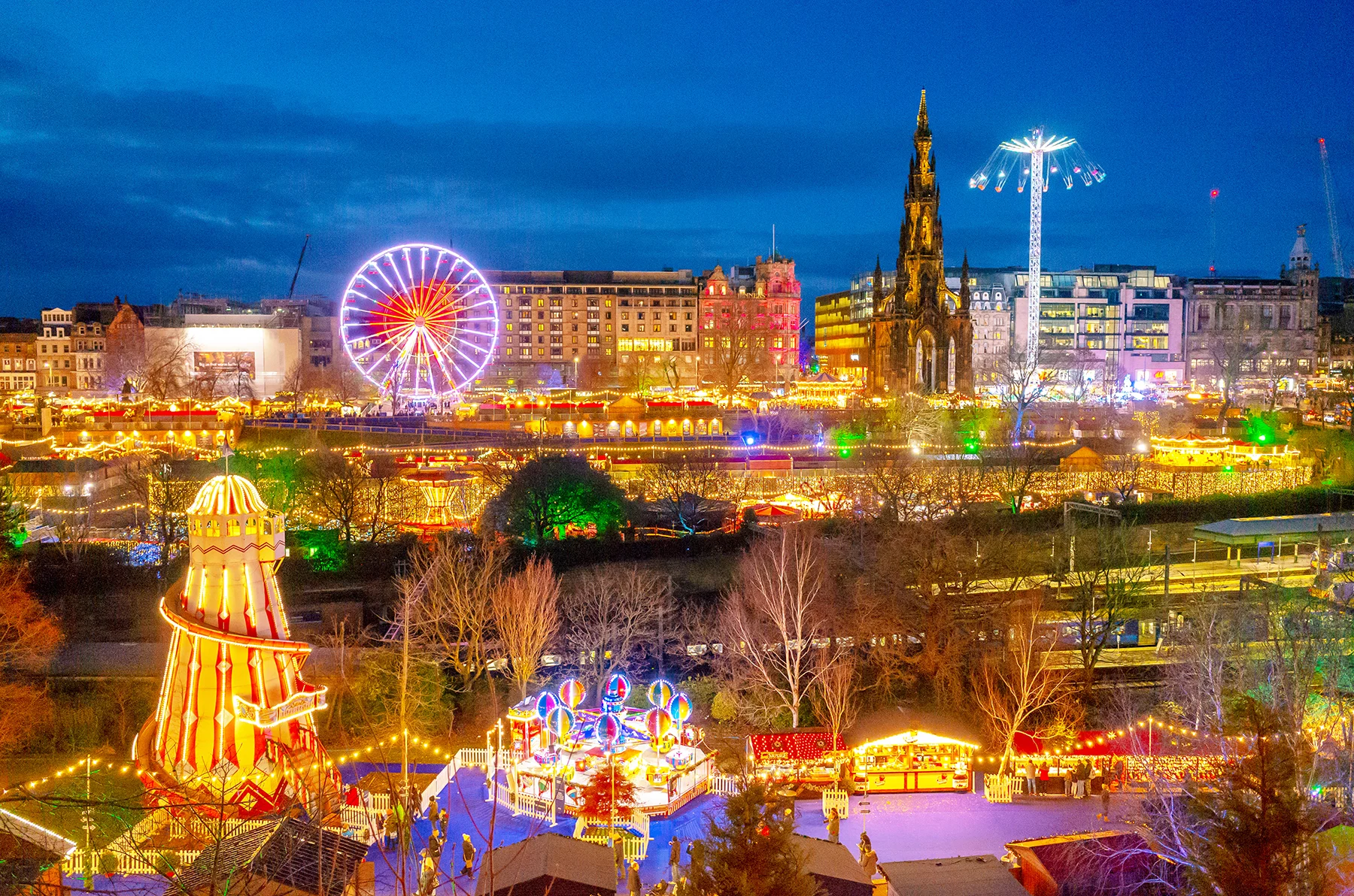A Christmas fair at night, with bright multicoloured lights and a view across Edinburgh city centre.