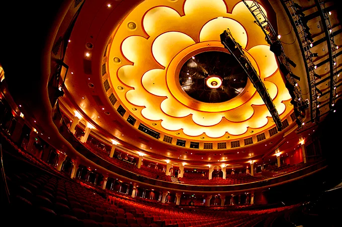 Looking up from the auditorium of a theatre with red velvet seats to an elaborate domed ceiling with ornate white and gold mouldings.