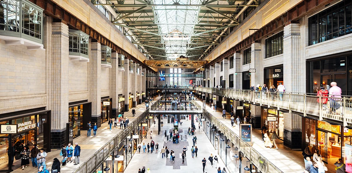 The interior of a large shopping centre with wide walkways and multiple levels. People are walking through the centre and there is a high glass roof above. 