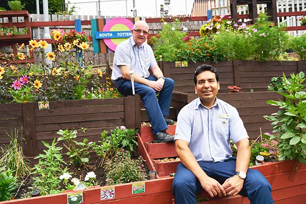 2 men in railway uniforms sitting on planters for a colourful garden at a train station. 