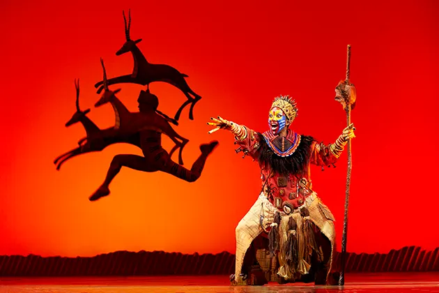 A performer in African dress and make-up onstage at The Lion King, against a red backdrop with silhouettes of gazelles.