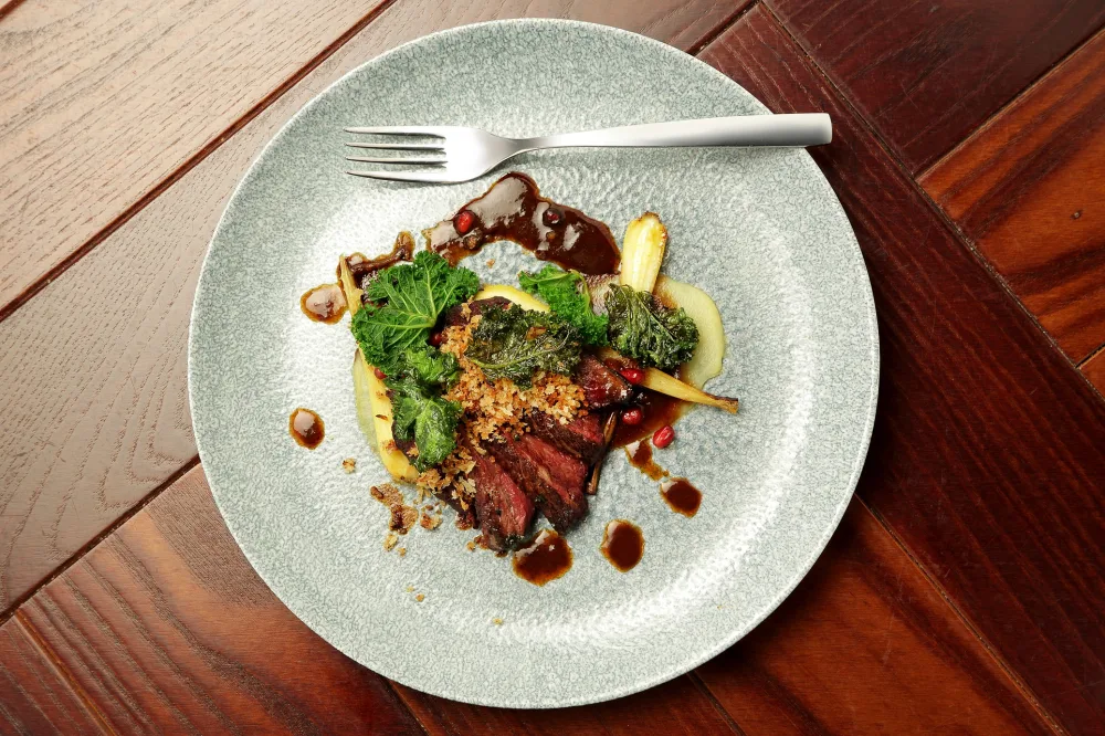 A grey plate on a wooden table with a metal fork, the dish contains sliced red meat, gravy and vegetables elegantly arranged.