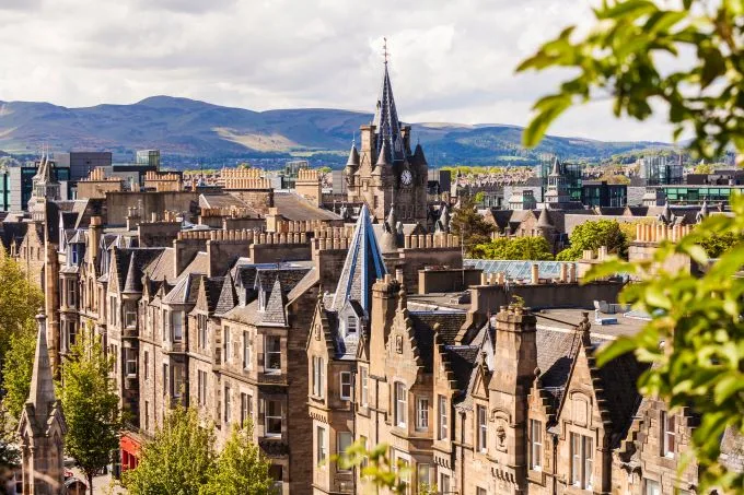 The jagged skyline of Edinburgh's iconic buildings in creams and browns, with mountains in the background and green foliage framing the image.