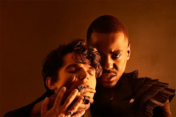 A Black actor (Ncuti Gatwa) stands behind a white actor (Edward Bluebell), holding his hand across his mouth. 