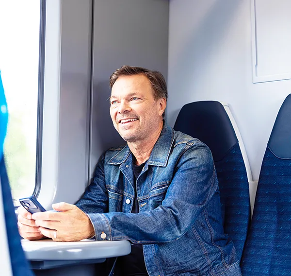 Pete Tong, a smiling middle-aged man in a denim jacket, holding a smartphone and seated by a window on a train.