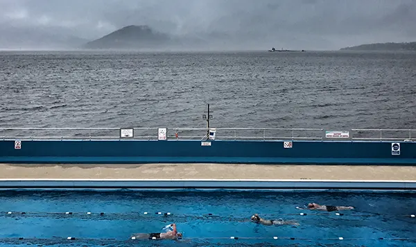 Swimmers in an outdoor pool on a grey, overcast day, with a calm sea and misty hills visible in the background.