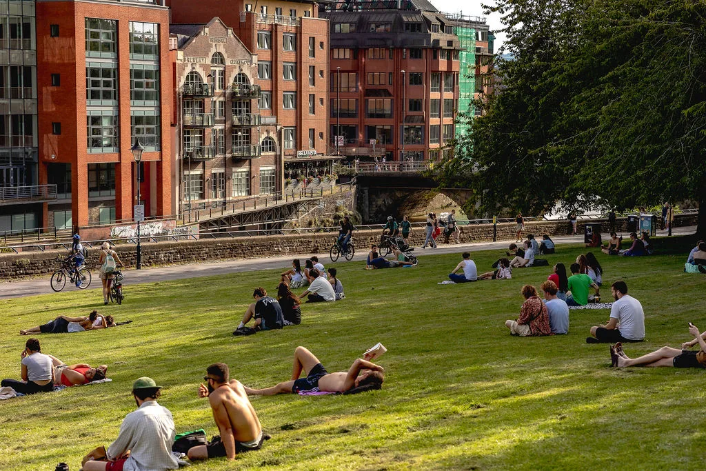 Groups of people relaxing and sunbathing on a green lawn next to a canal. 
