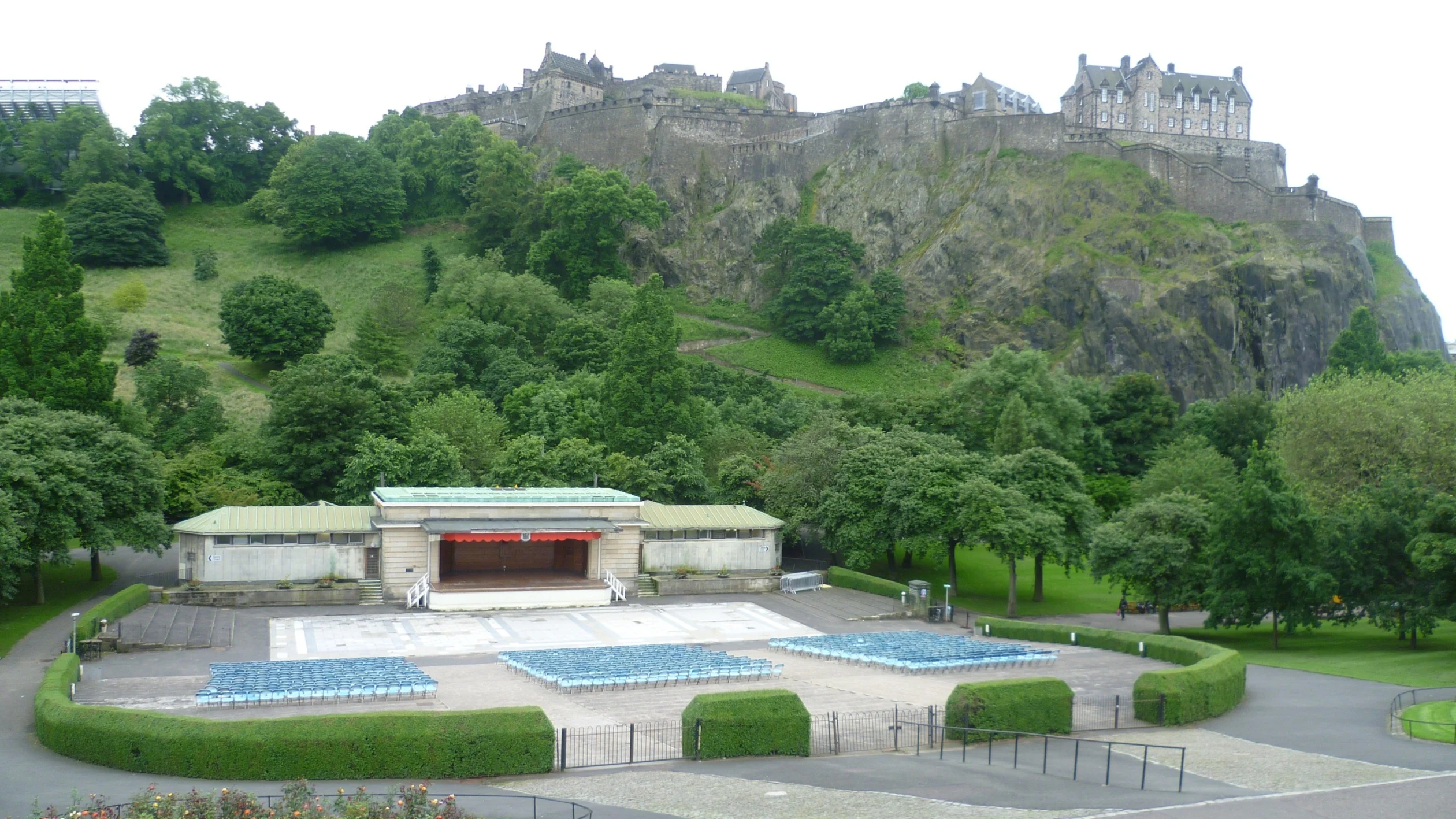 An empty open air bandstand with lots of seats, sitting in front of a large green hill with a castle on the top. 