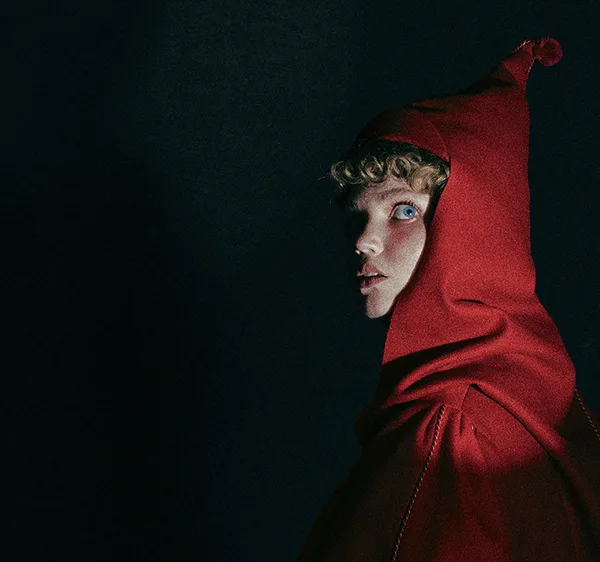 A young white woman with blue eyes looking scared, wearing a red cape and cap, against a black background.