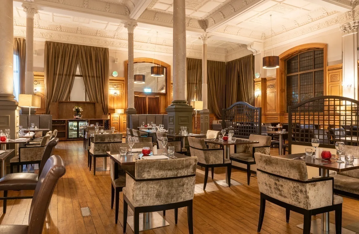 An empty hotel dining room with grey velvet chairs and black tables, there are large white columns and high ceilings, with beige curtains draped at the windows.