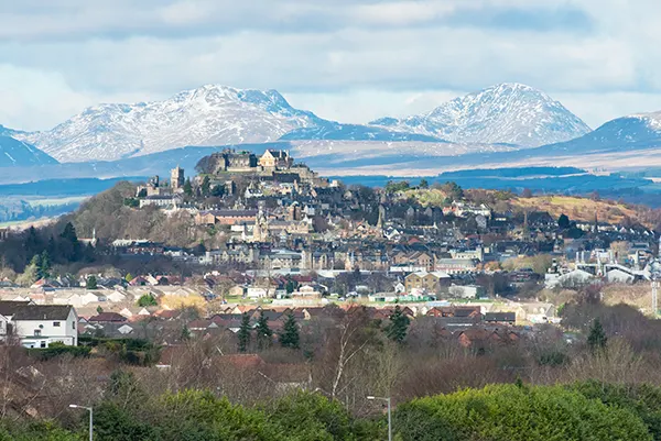 A castle sitting on a hill above a small town, with mountains in the background. 