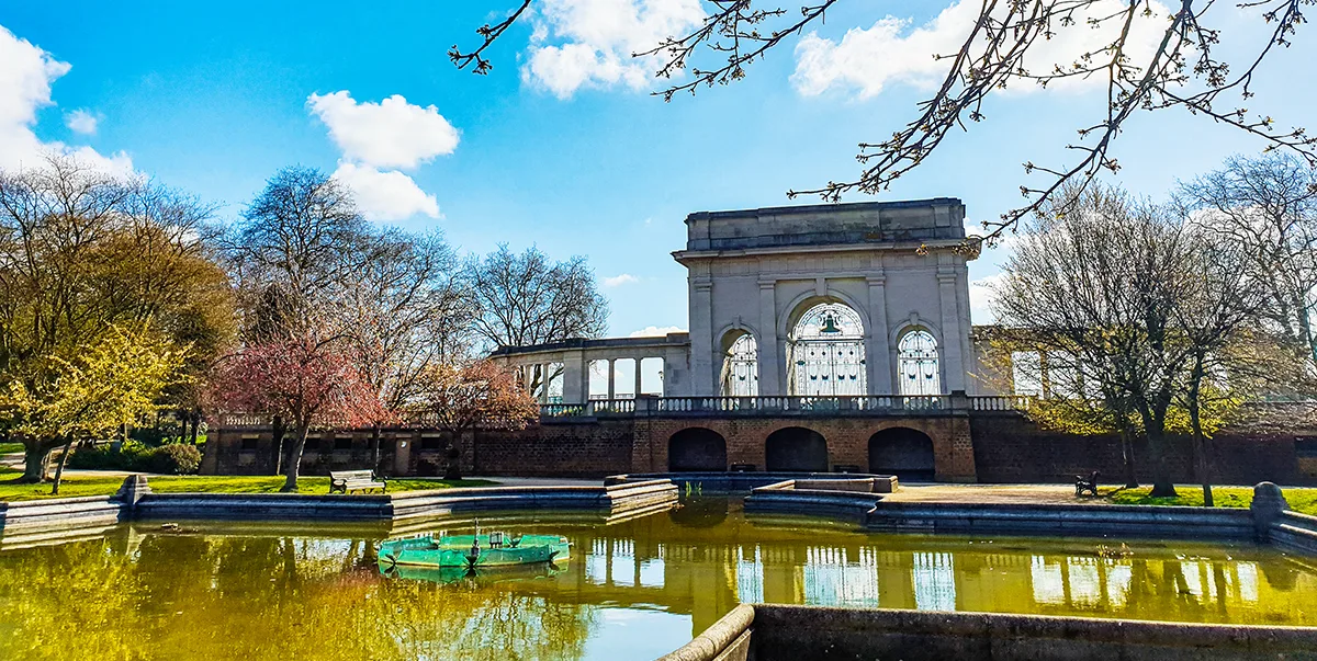 A white stone war memorial with 3 arches alongside a park and a pond. 