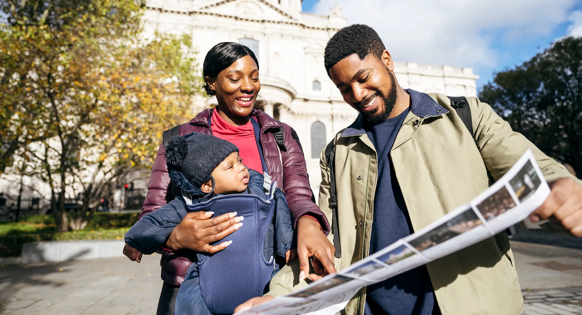 A Black woman who is wearing a baby in a sling stands next to a Black man, they are both looking at a fold-out leaflet and smiling.