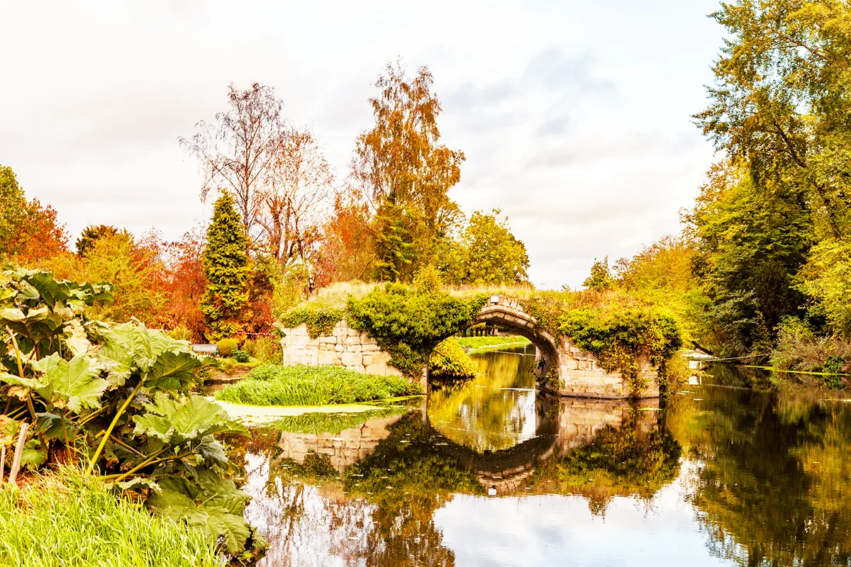 Stone bridge over calm water surrounded by autumn trees with vibrant orange, yellow and green foliage reflecting in the water.