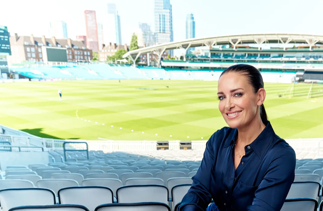 Kirsty Gallacher, a white woman with long dark hair pulled back in a ponytail, sitting in an empty sports stadium, she is smiling and looking at the camera.